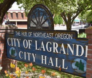 Blue City of La Grande sign in front of historic City Hall with flowers blooming 
